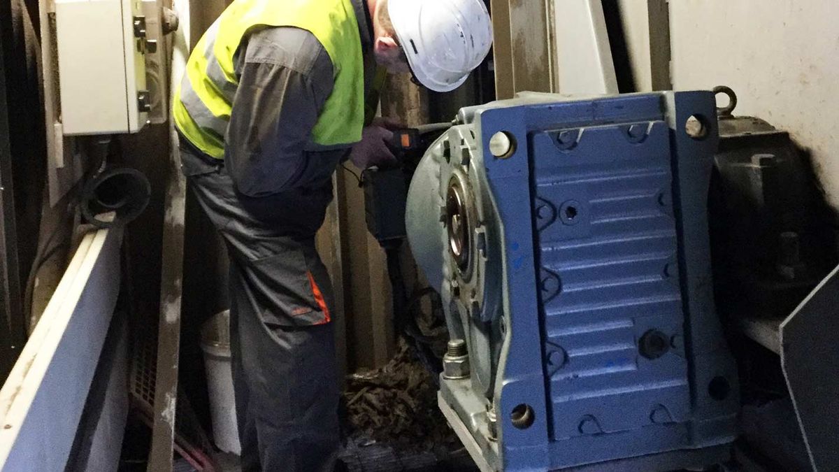 Technicus met helm en geel hesje boort naast een blauwe motorreductor in een installatie