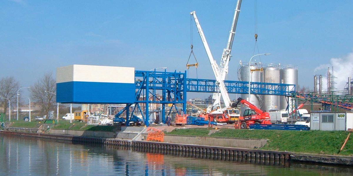 Grue levant une galerie de convoyeur bleue sur un site industriel au bord d'un canal