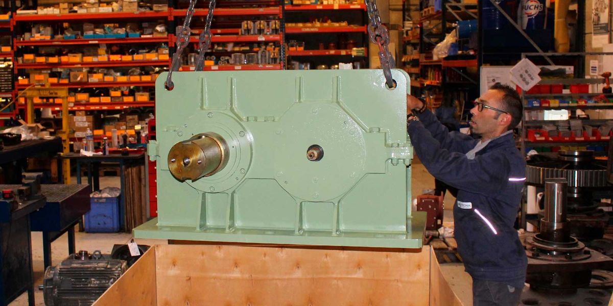 Technicien guidant un réducteur industriel vert levé à la chaîne au-dessus d'une caisse en bois, dans l'atelier