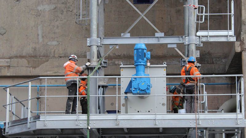 Techniciens en tenue orange posant un réducteur bleu sur une plateforme métallique en hauteur