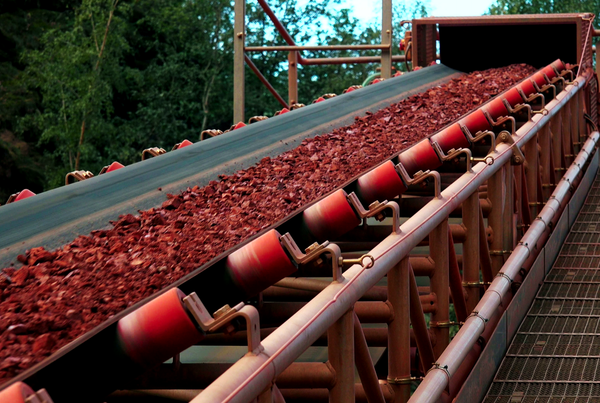 Convoyeur à bande incliné chargé de minerai rouge, rouleaux porteurs rouges et passerelle grillagée