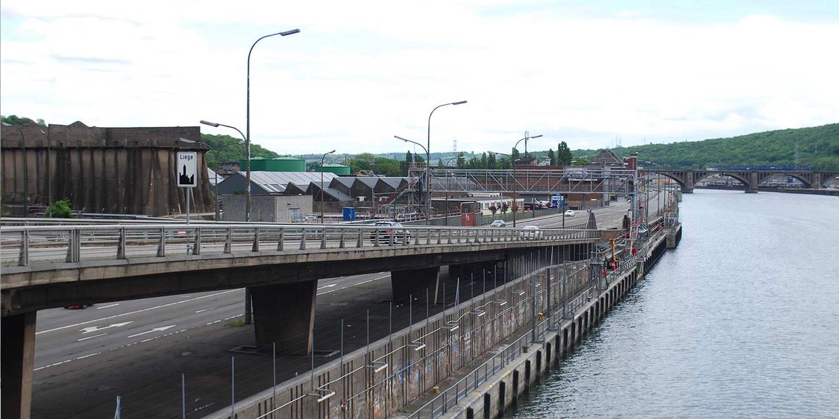 Autoroute urbaine surélevée le long d'un fleuve, panneau d'entrée de Liège, collines et pont au loin