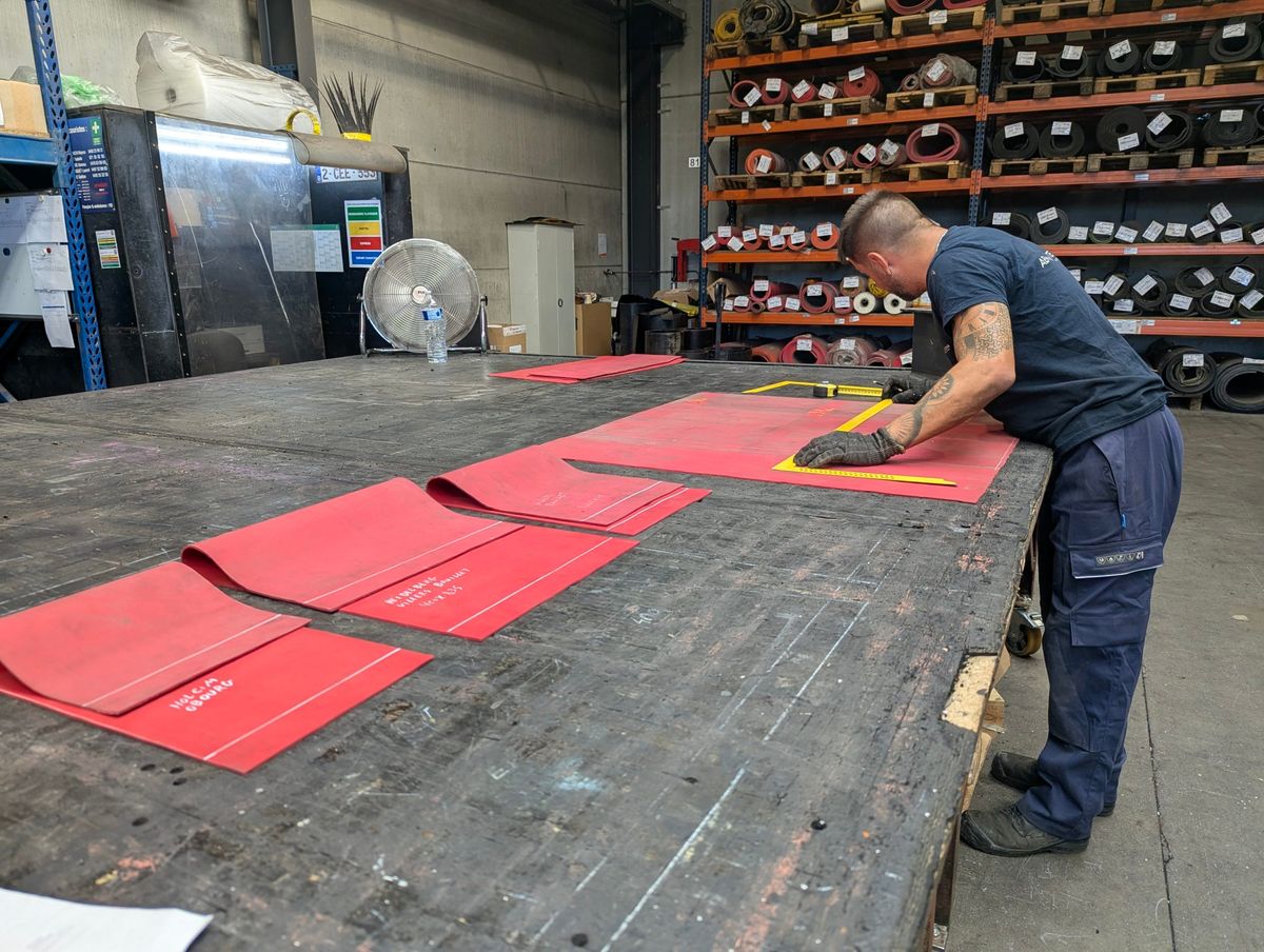 Technicien découpant des plaques de caoutchouc rouge sur la table de l'atelier, rouleaux de bande en rayonnage