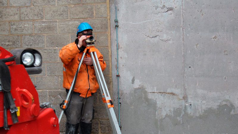 Technicien casqué relevant des mesures sur site avec un niveau optique sur trépied, devant un mur en béton