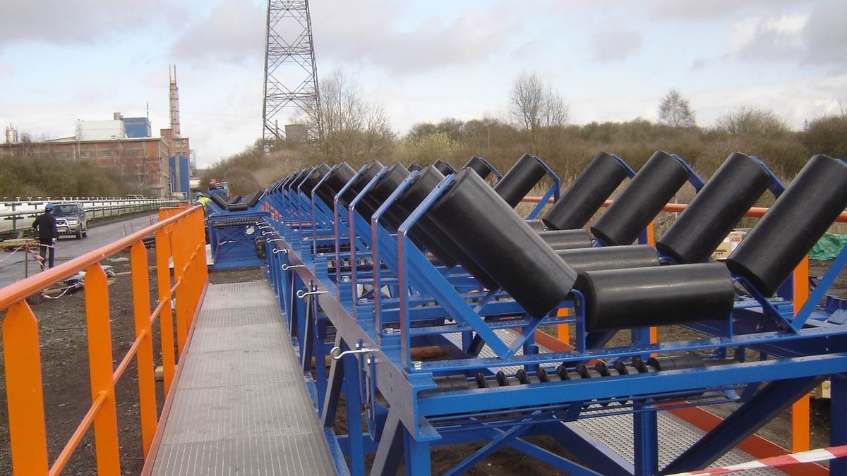 Buitenopgestelde transportband: troggen met zwarte rollen op blauw frame, loopbrug met oranje leuning
