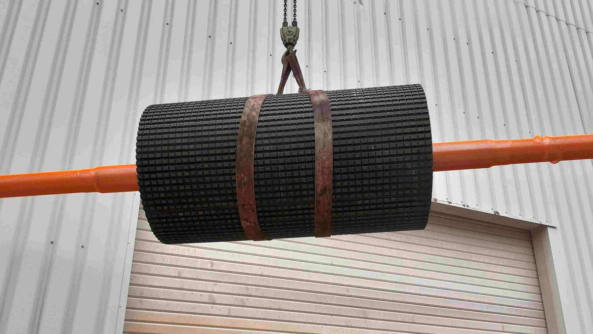 Transportbandtrommel met gegroefde bekleding aan een kraan hangend, oranje as, voor een metalen gevelbeplating