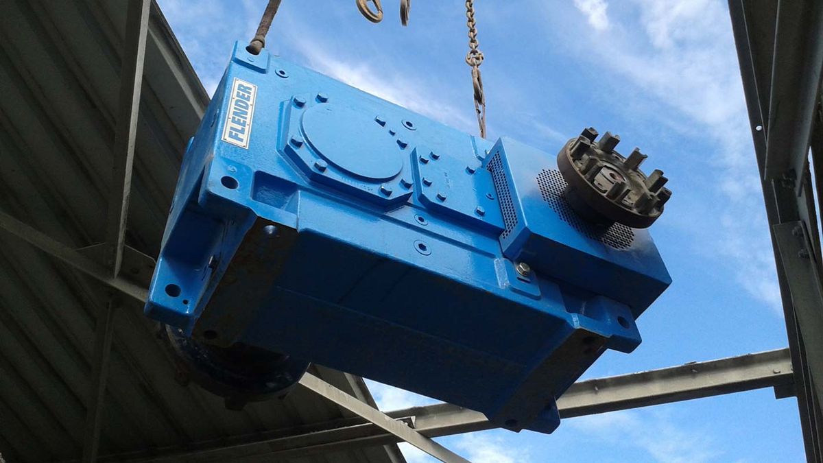 Réducteur Flender bleu suspendu à une chaîne de levage sous une charpente métallique, ciel bleu en arrière-plan