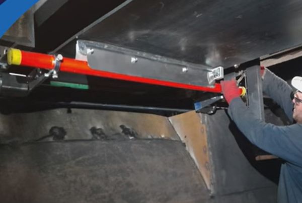 Technicien posant un racleur à bras rouge sous une bande, près d'une trémie